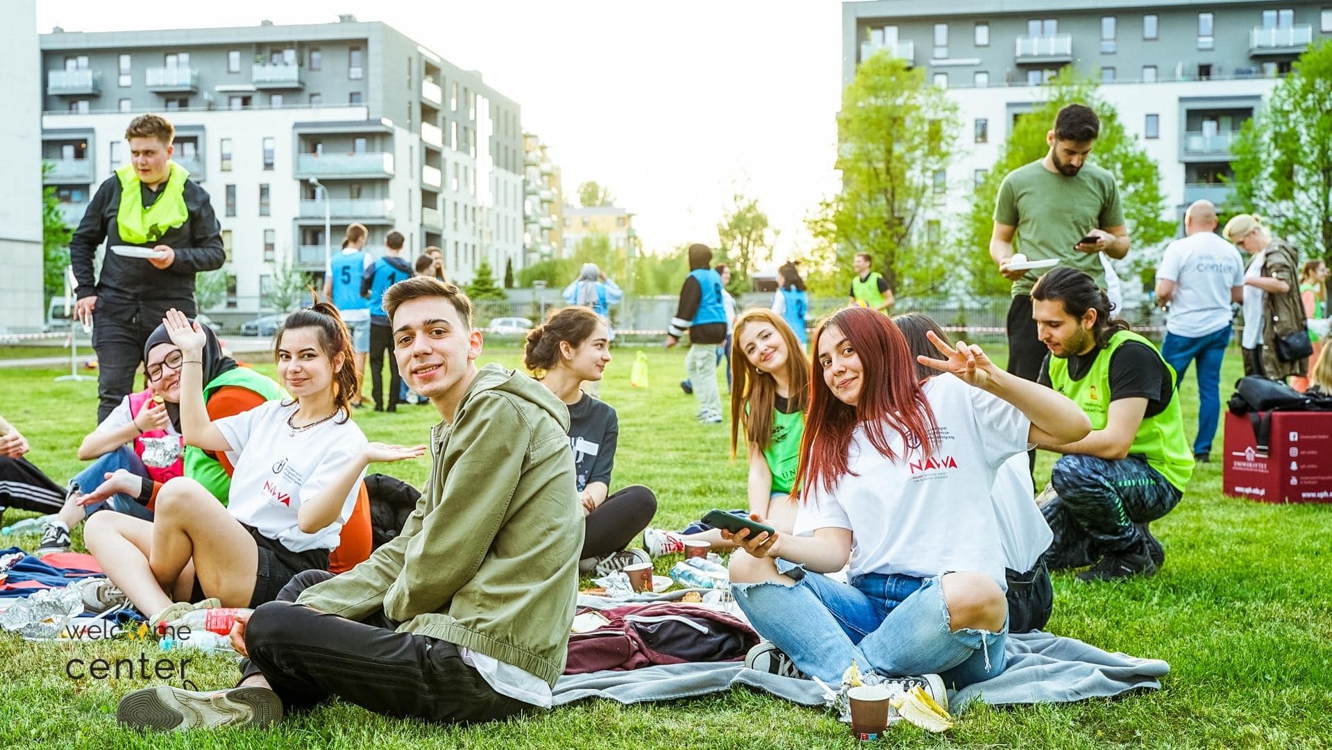 Studenci Erasmus+ wspólnie uczestniczą w aktywnościach sportowych na świeżym powietrzu. Studenci siedzą na kocykach na trawie i pozują do zdjęcia.